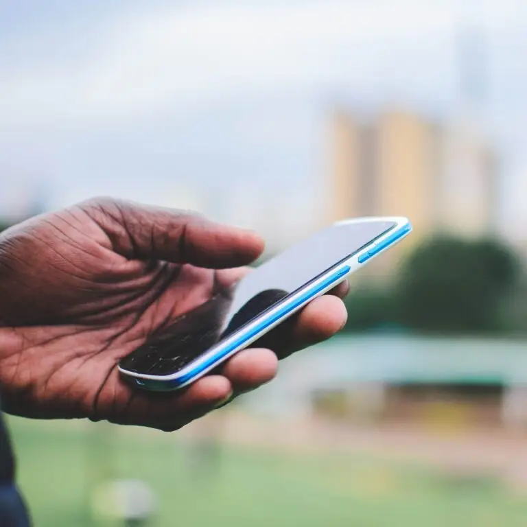 A close-up of a hand holding a smartphone outdoors with blurred urban background.